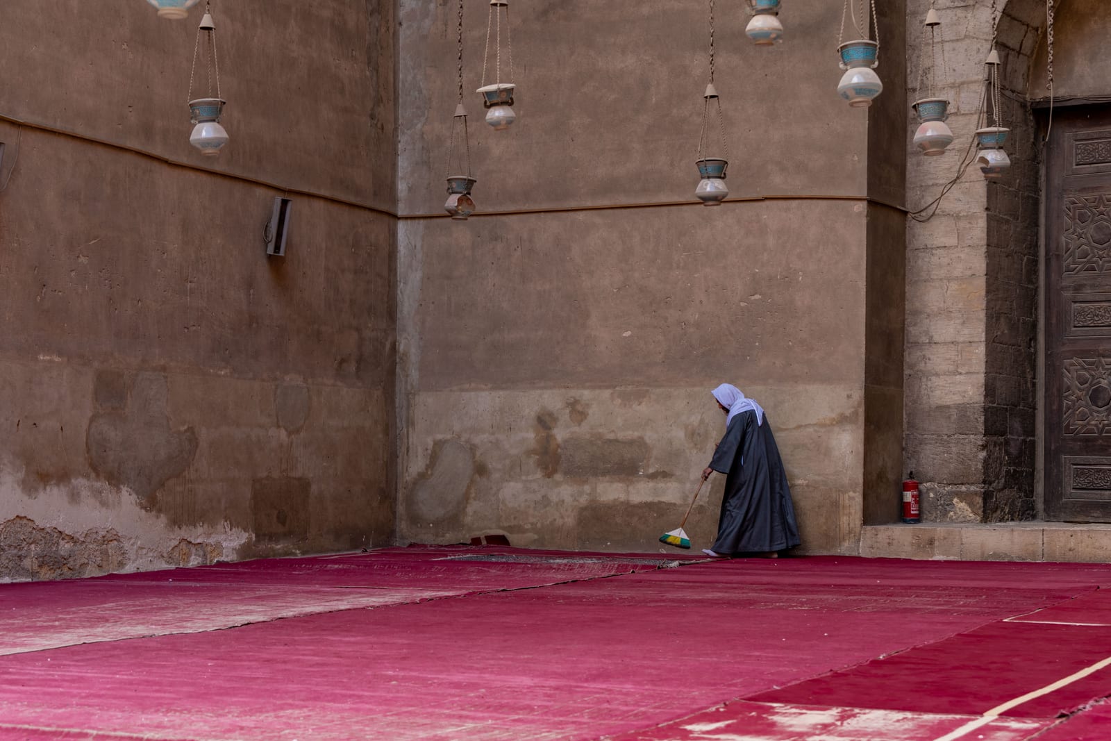 A caretaker sweeps the red carpets of the vast Mosque–Madrasa of Sultan Ḥasan in Cairo, light falling across the stone walls.