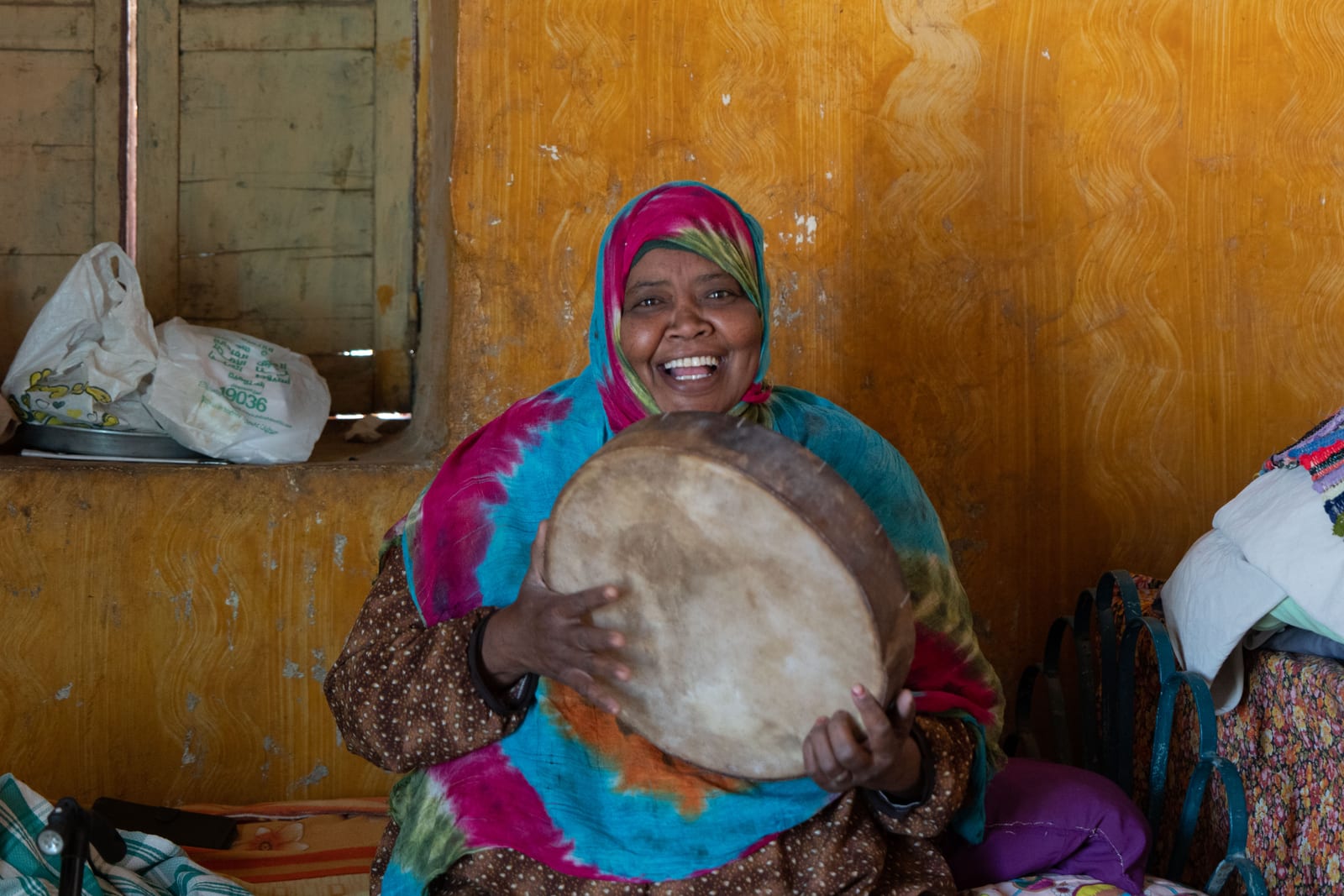 A Nubian woman in a bright headscarf laughing as she plays a frame drum in front of an ochre wall.