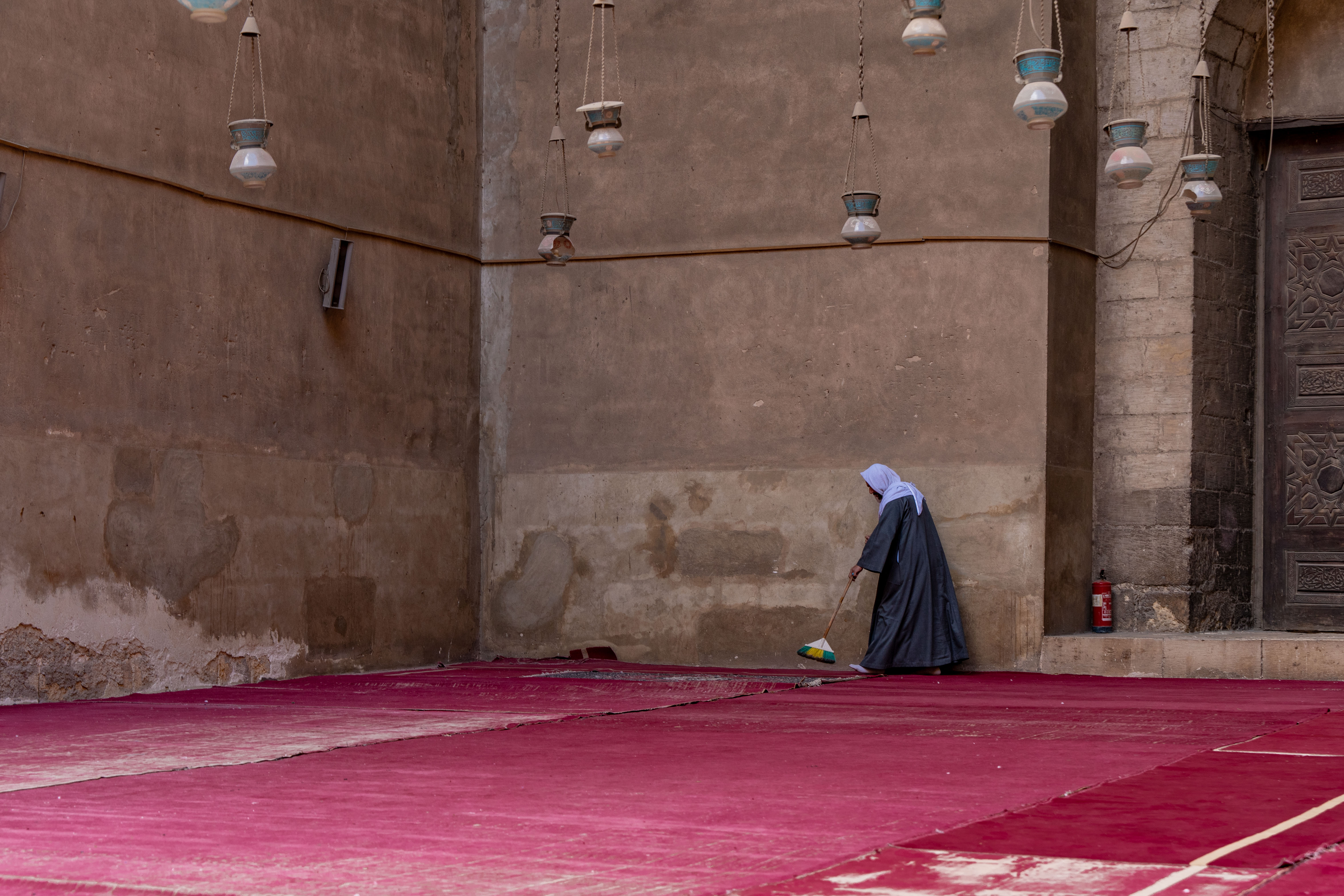 “Cinderello” — a caretaker sweeping patterned carpets inside the Mosque–Madrasa of Sultan Hasan, sunlight cutting across the vast stone interior.