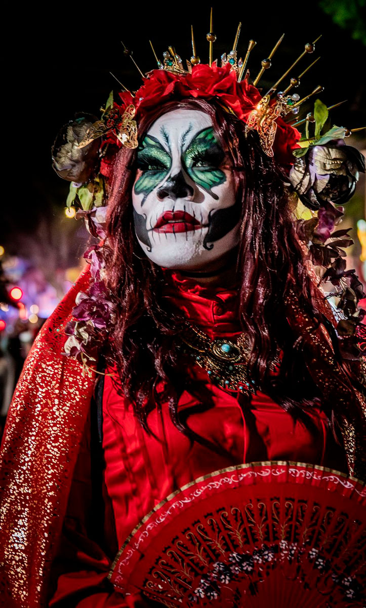 A woman in Day of the Dead calavera face paint and a crown of red roses, holding a red fan at night in the Mission District, San Francisco.