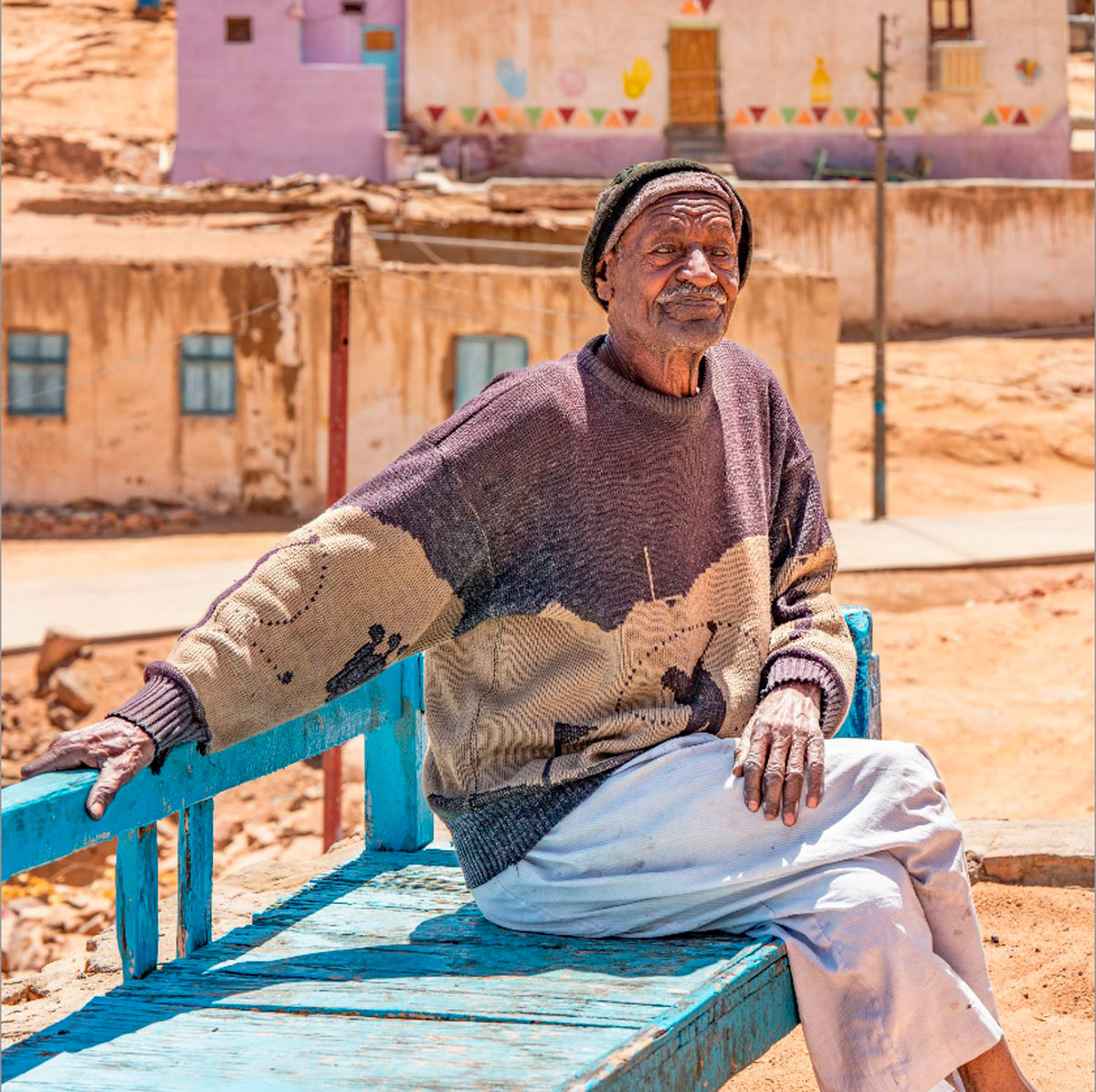 An elderly man resting on a blue bench in a Nubian village, gazing upward.