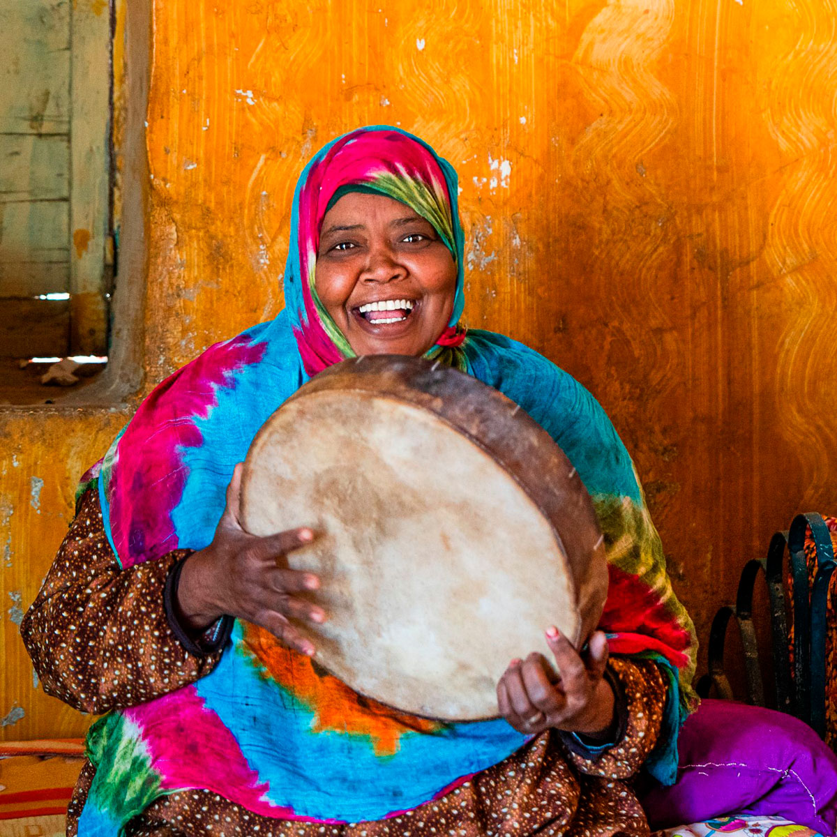 A Nubian woman in a tie-dyed headscarf laughing as she plays a frame drum.