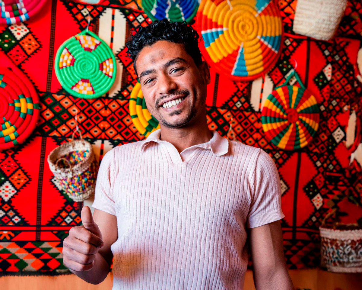 A smiling craftsman giving a thumbs up in front of colorful woven baskets.
