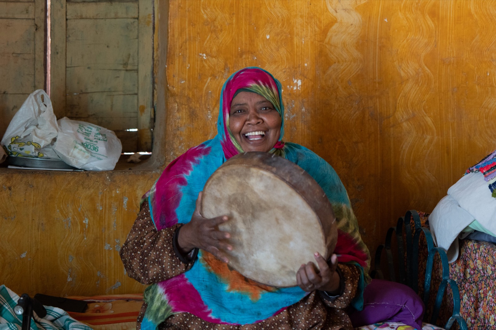 A Nubian woman in a bright tie-dyed headscarf laughing as she plays a frame drum in front of an ochre wall.