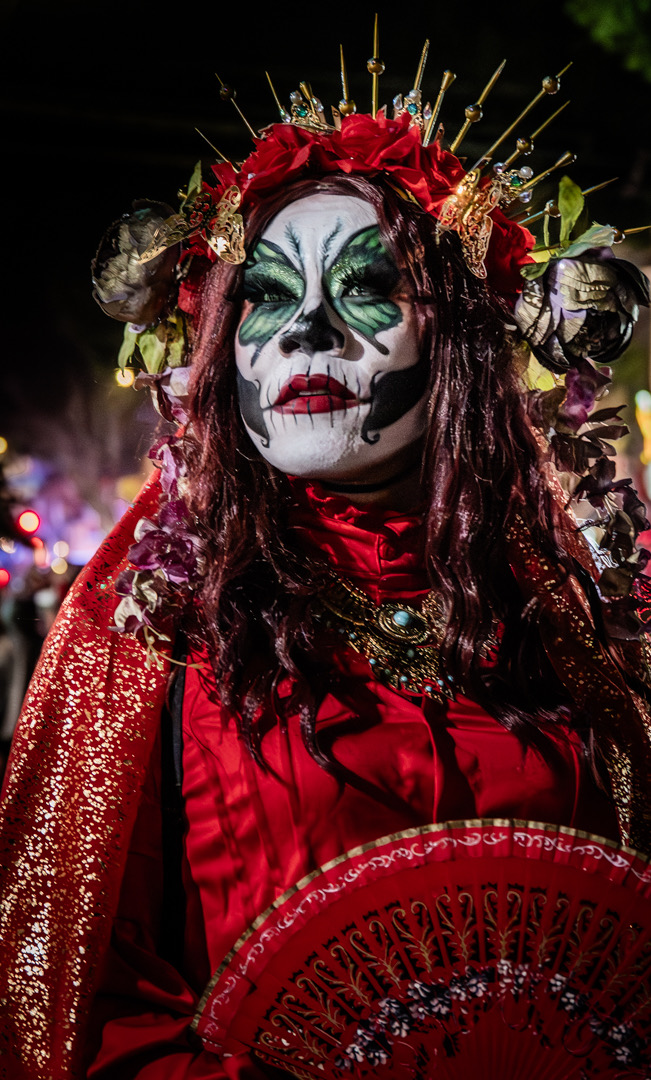A woman in a red dress with Day of the Dead calavera face paint, a crown of red roses and gold spikes, holding a red fan at night in the Mission District, San Francisco.